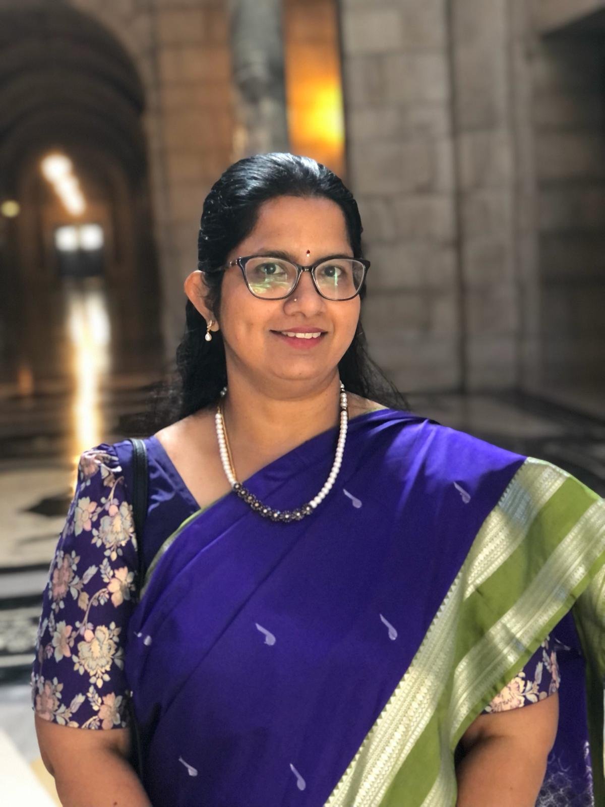 A woman wearing glasses, a purple and green traditional Indian sari, a pearl necklace, and earrings, standing in a corridor with stone walls and warm lighting