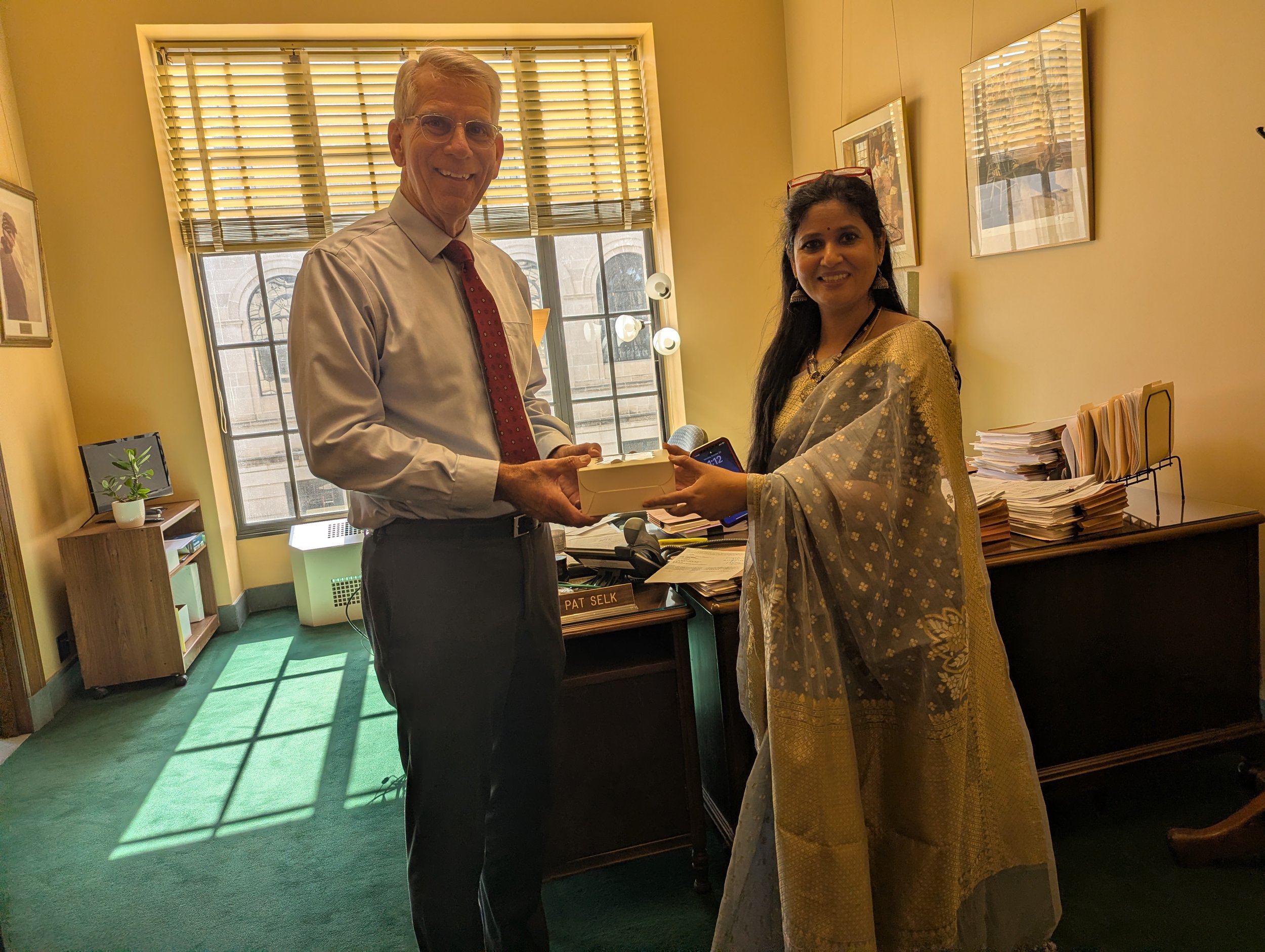 A man in a suit and tie hands a woman in traditional Indian attire a small box. They are in an office with a large window, framed pictures, and a desk with stacks of papers.