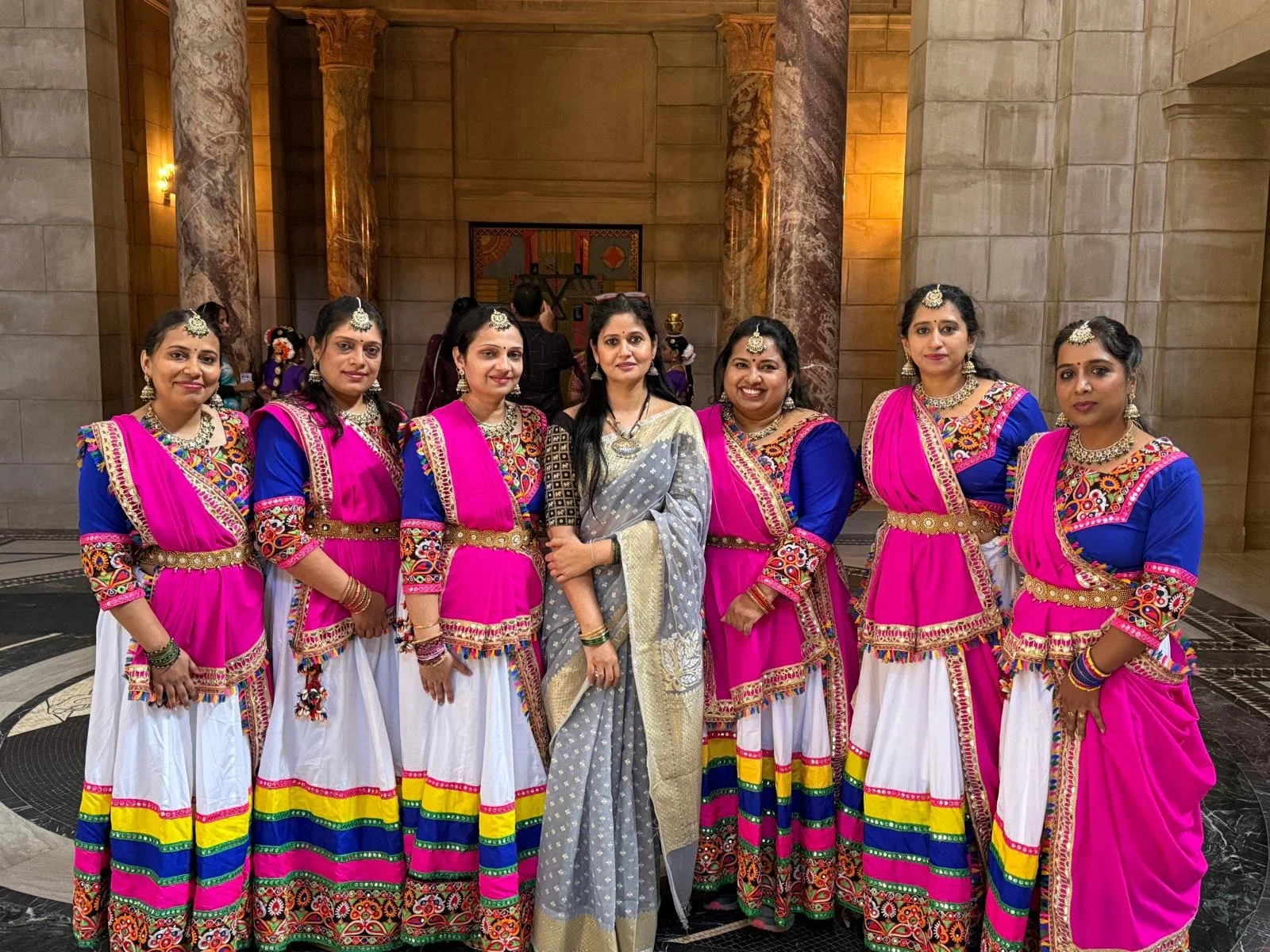 Group of seven women dressed in traditional Indian attire, with five wearing blue and pink dresses with intricate embroidery, and two in grey and gold sarees, standing inside a grand hall with marble columns and warm lighting.