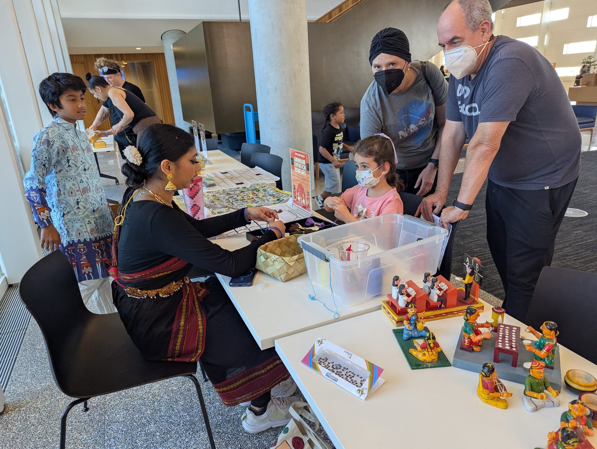 People at a table displaying traditional Indian dolls and crafts, with children and adults engaging and browsing the items, in an indoor setting with bright natural light.