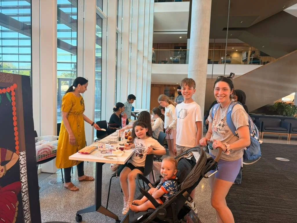 A group of children and adults gathered around a table inside a modern building with large windows. Some children are sitting, and one is in a stroller. Adults are standing and talking. The environment appears bright and welcoming, with a staircase a