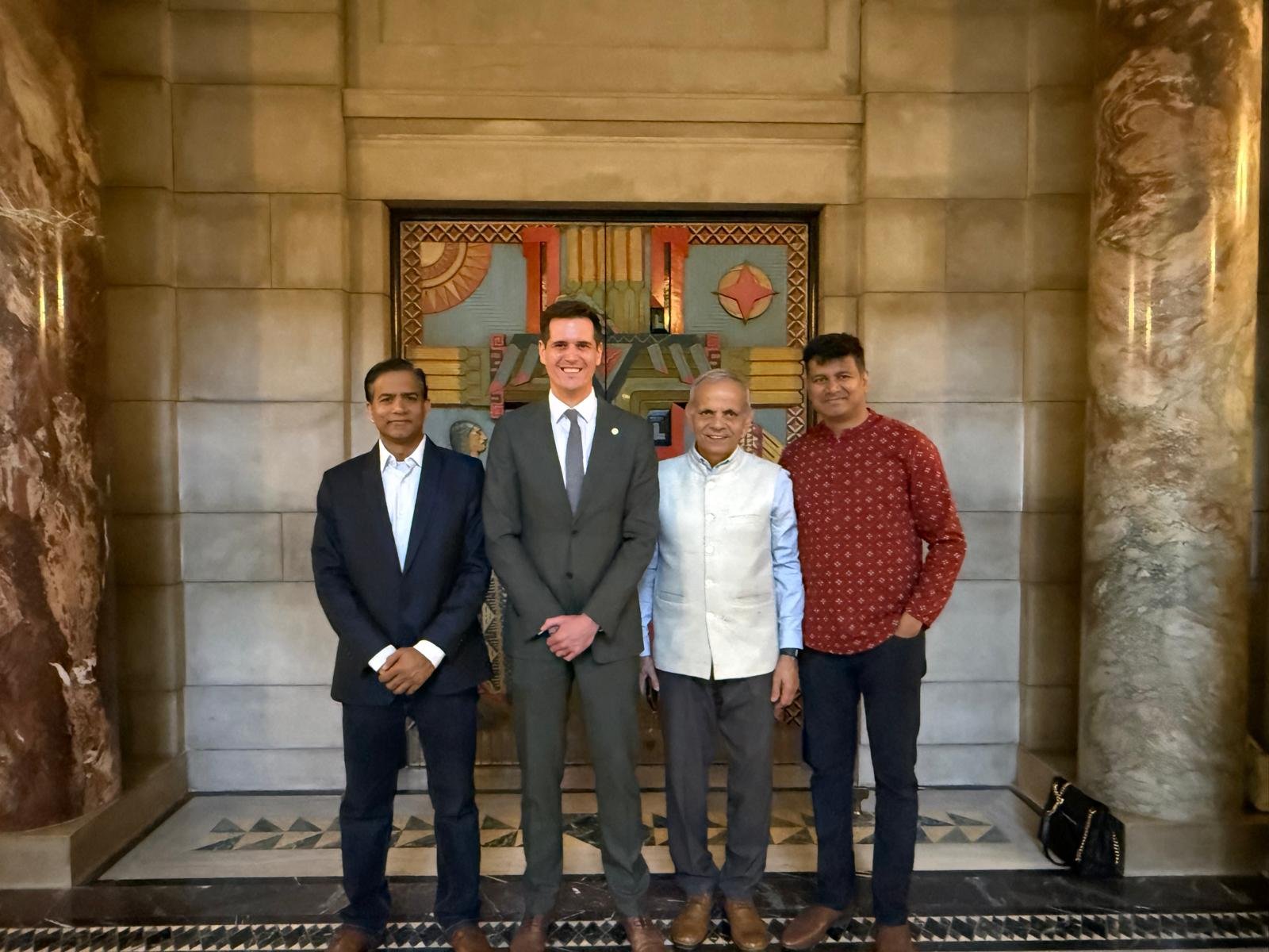 Four men standing in front of a colorful artwork on a stone wall. Three are dressed in formal attire, and one is in a patterned red shirt. The setting appears to be an elegant indoor location.