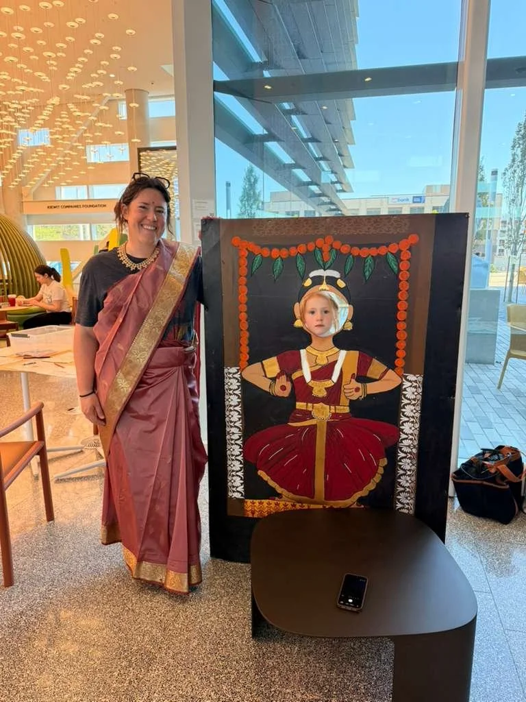 A woman dressed in traditional Indian attire standing next to a painted cutout of a girl in traditional Indian dance costume, inside a modern building with large glass windows.