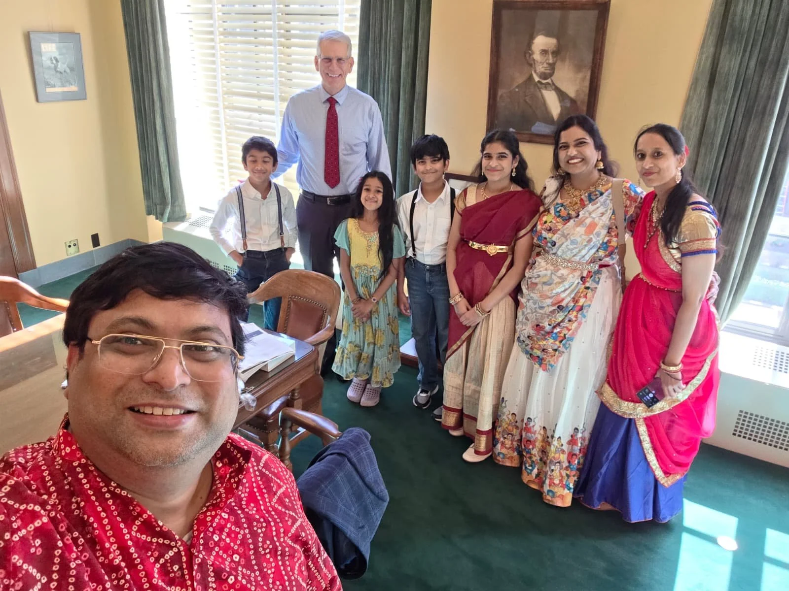 A group of people, including children and adults, standing in a room with large windows and green curtains. The person in the front is taking a selfie with the group. Several individuals are dressed in traditional Indian attire, while others are in c