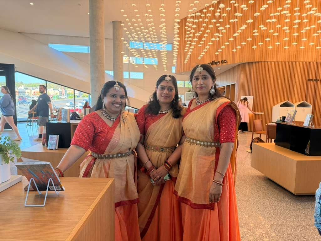 Three women dressed in traditional Indian sarees with jewelry, standing inside a modern, well-lit building with wooden and light fixtures on the ceiling.