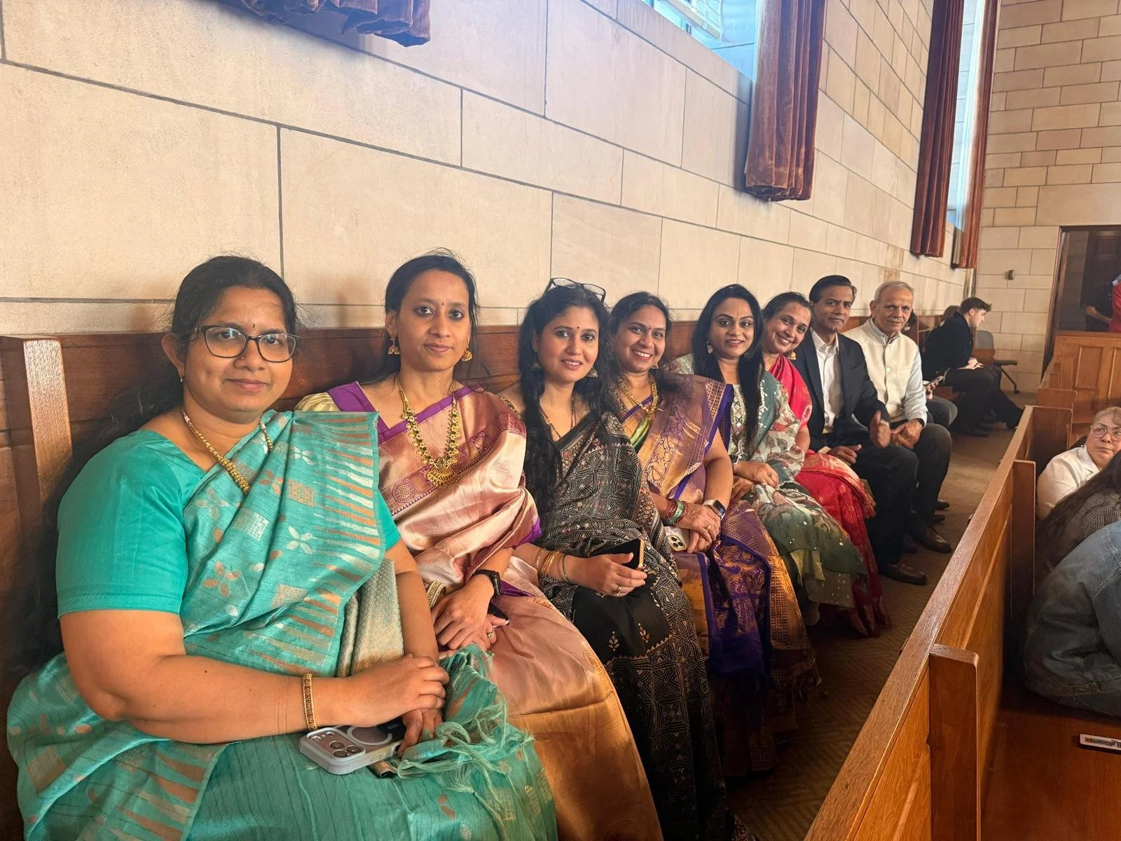 A group of women and men sitting on wooden pews in a church, dressed in traditional Indian attire, smiling for the photo.