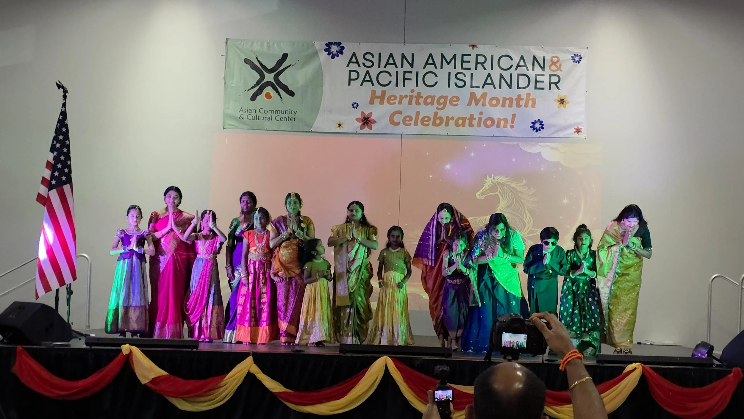 Group of children and adults dressed in colorful traditional Indian attire performing on stage during an Asian American and Pacific Islander Heritage Month celebration, with a banner and U.S. flag in the background.