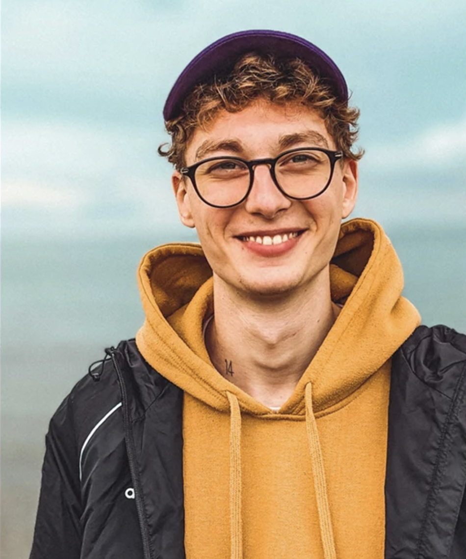 A young man smiling outdoors, wearing glasses, a purple cap, a mustard yellow hoodie, and a black jacket, with a cloudy sky in the background.