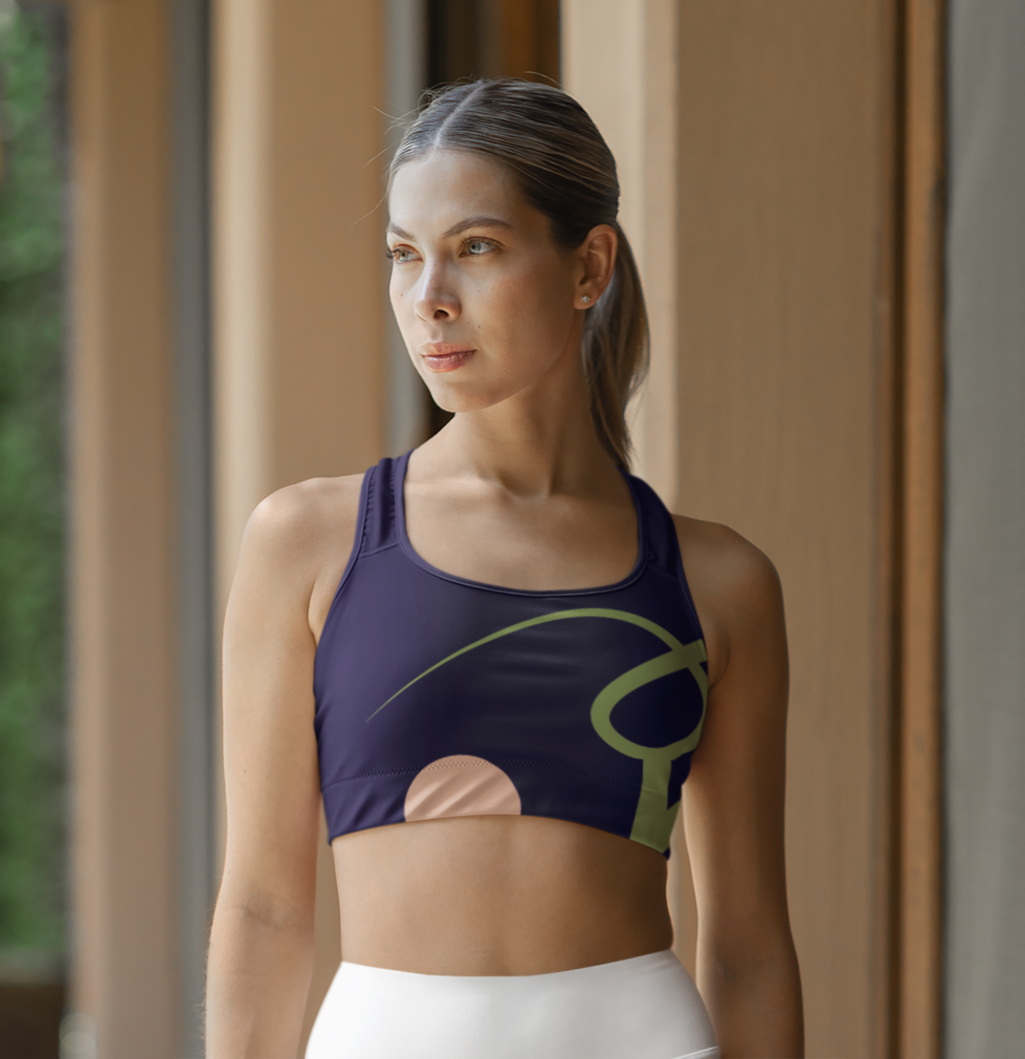 A woman with long brown hair tied back, wearing a dark purple tank top with a yellow logo, standing indoors near a window with a blurred background.