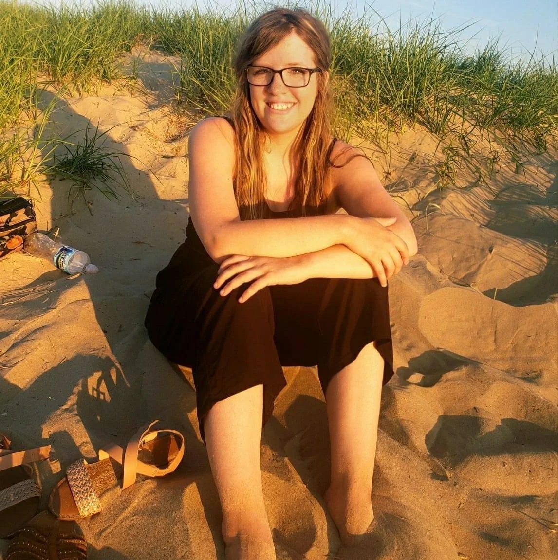 A woman with glasses, long hair, wearing a black dress, sitting on a sandy beach with grass and dunes in the background, smiling at the camera during sunset.