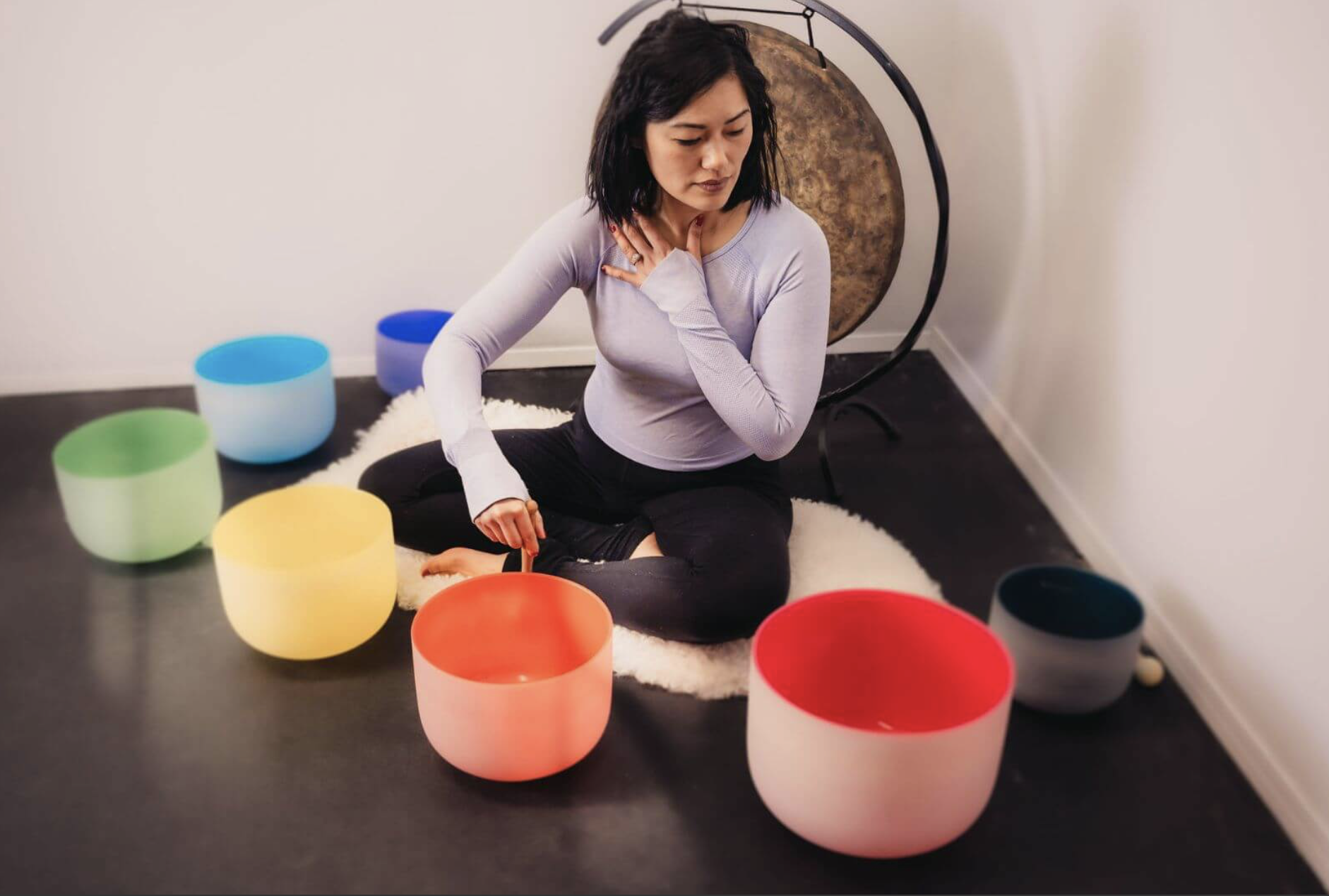 A woman practicing yoga or meditation while sitting cross-legged on a white rug, with her hand on her chest and her eyes closed. She is surrounded by colorful singing bowls on a dark floor in a minimalistic room with a plain white wall and a large gong in the background.