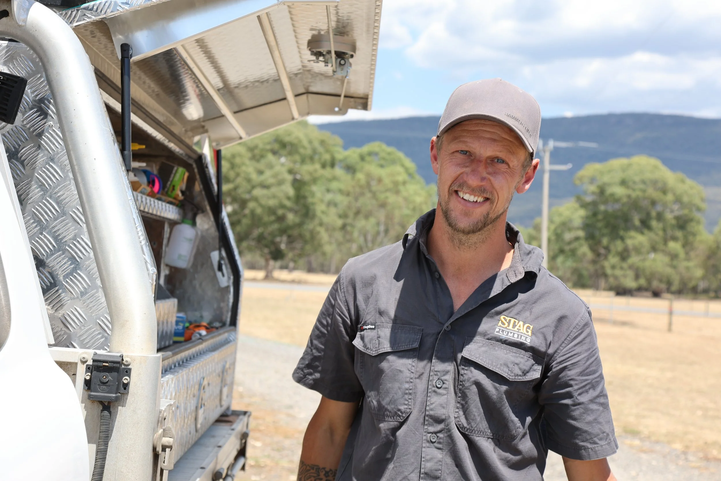 A smiling man wearing a gray cap and gray work shirt with a patch that reads 'STAG Plumbing' standing outdoors next to a plumbing service truck with an open side compartment filled with tools and supplies. Trees and hills are visible in the background.