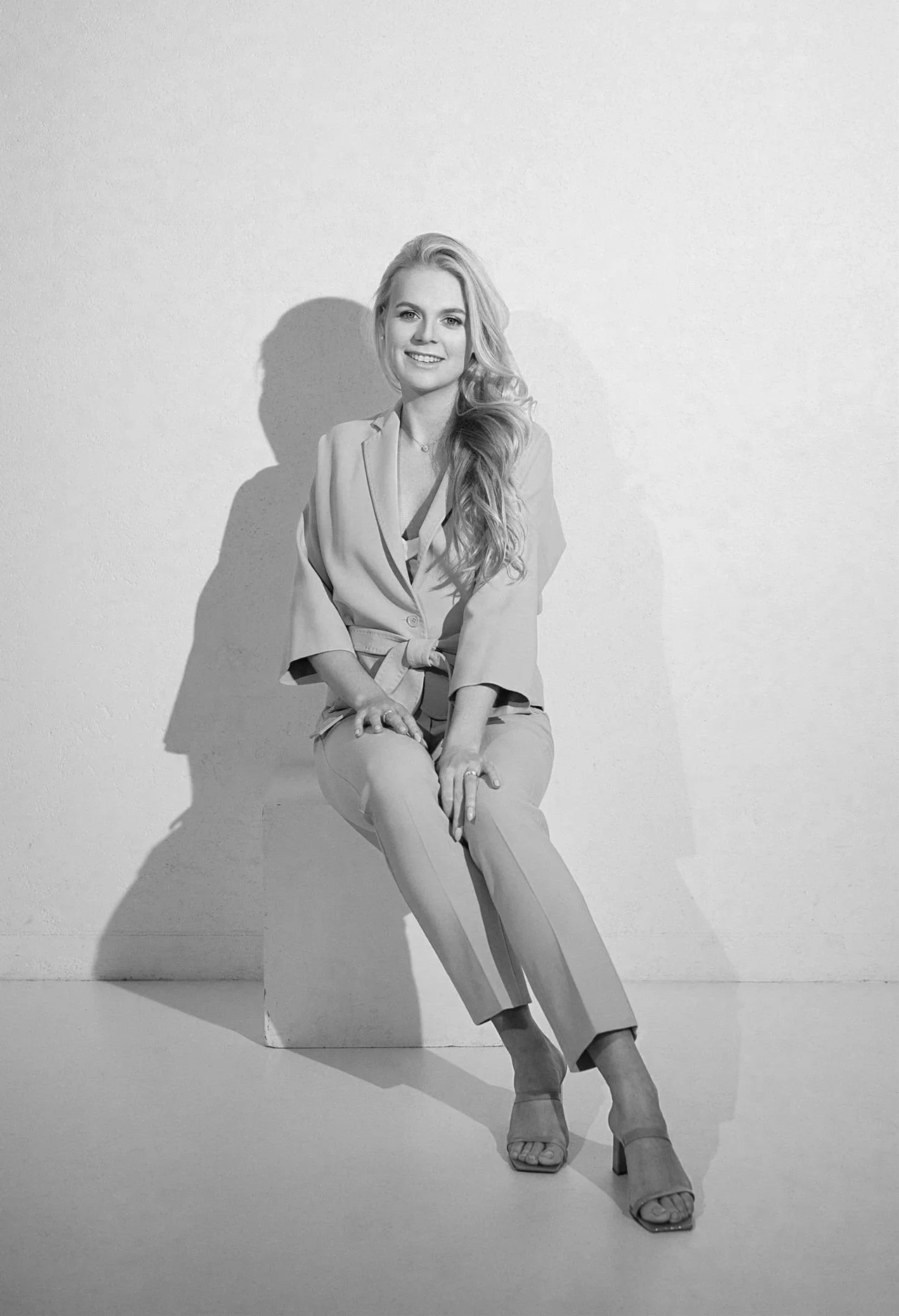 Black and white photograph of a woman sitting on a block against a plain wall, smiling, with long, wavy hair and wearing a blazer, trousers, and high-heeled sandals.