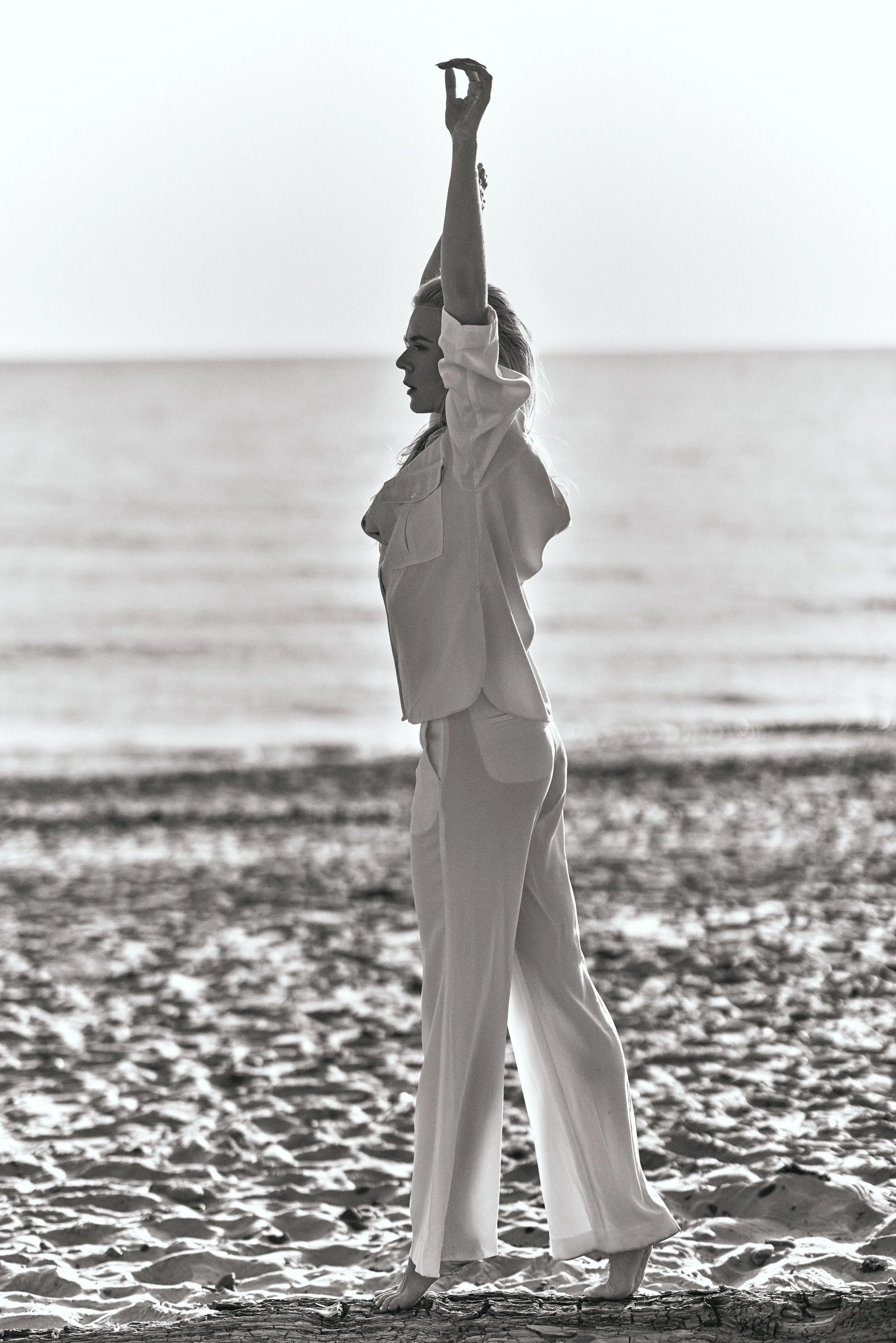 A woman in white clothing practicing yoga on a beach, standing on a log with ocean waves in the background.