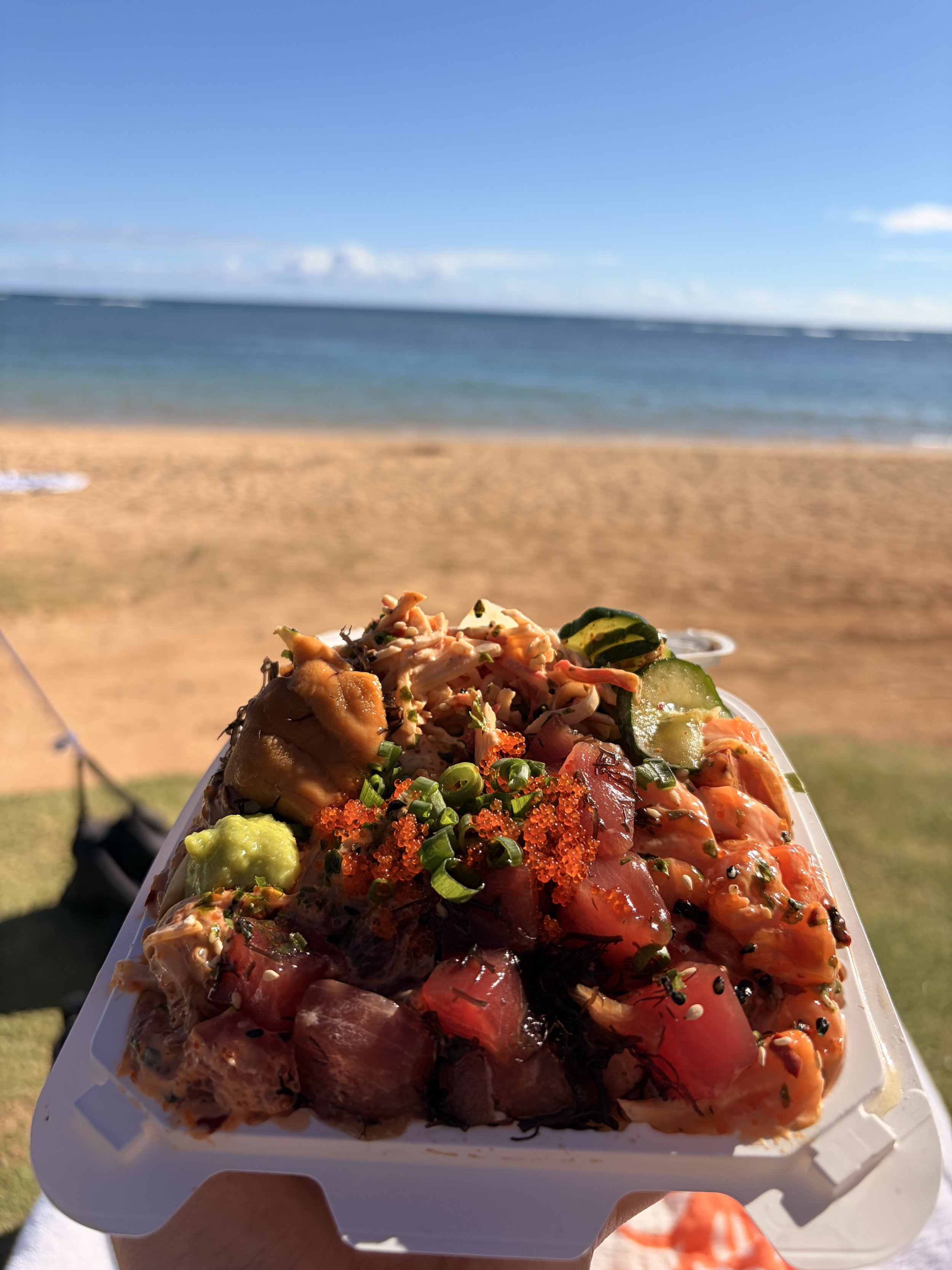 Close-up of a mixed seafood and vegetable poke bowl with salmon, cucumber, and fish roe, held in front of a beach scene with sand, ocean, and blue sky in the background.