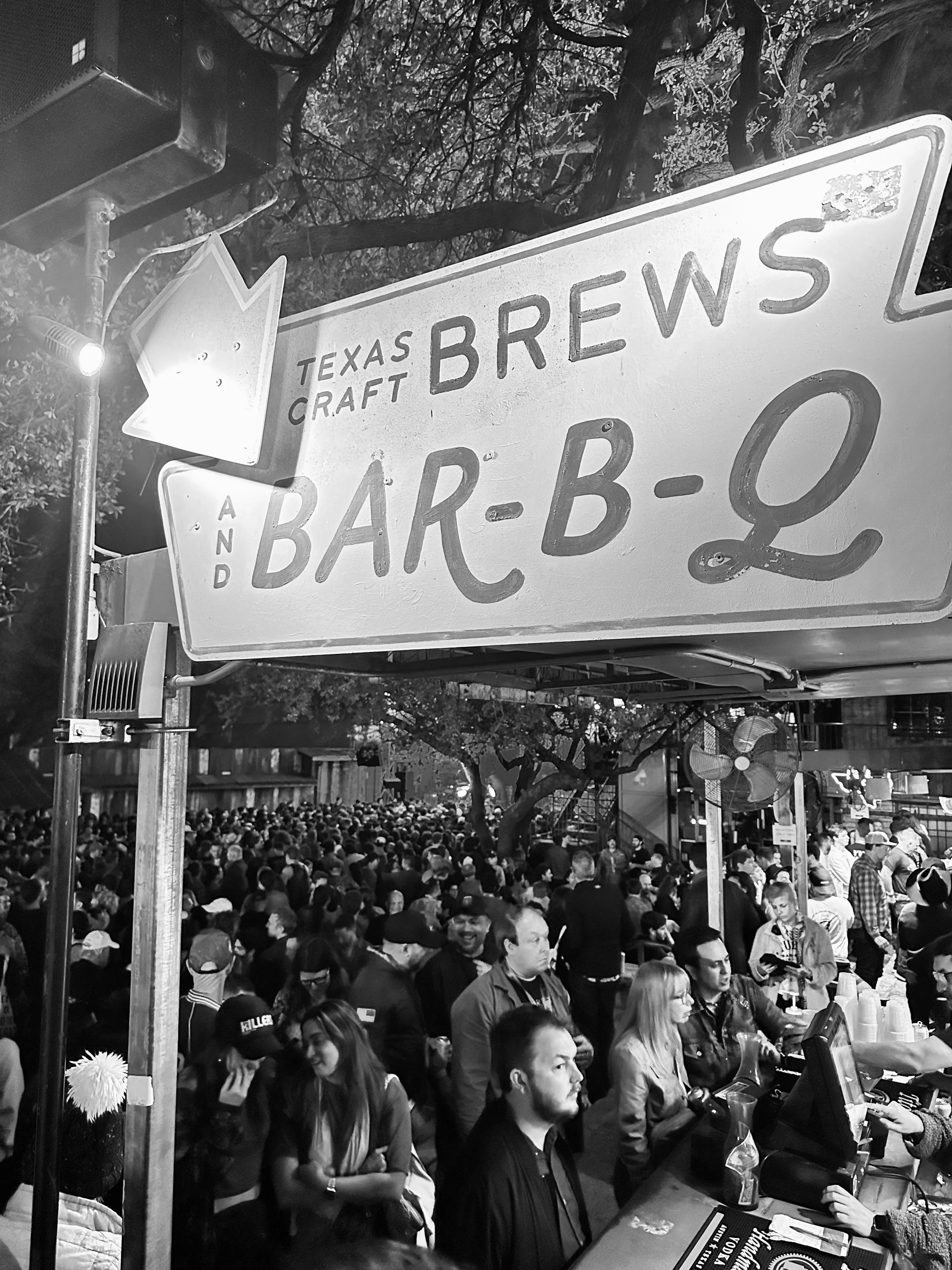 Crowd of people gathered outside at night, with a sign reading 'Texas Craft Brew Bar-B-Q' visible in the foreground.