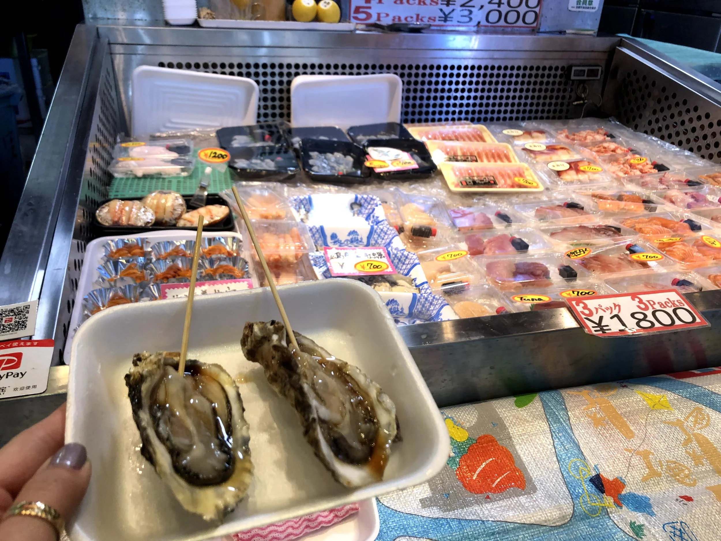 A hand holding a small white plate with oysters topped with sauce in front of a seafood display at a market, with various packaged seafood and price tags in the background.