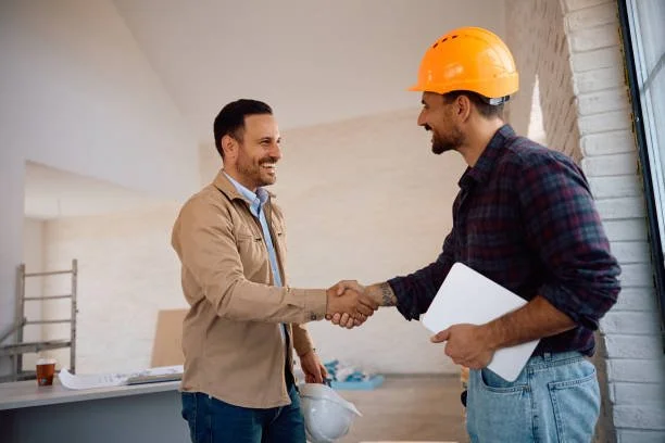 Two men shake hands inside a construction or renovation site, one in casual attire holding a hard hat, and the other wearing a plaid shirt and a yellow construction helmet, holding a clipboard.