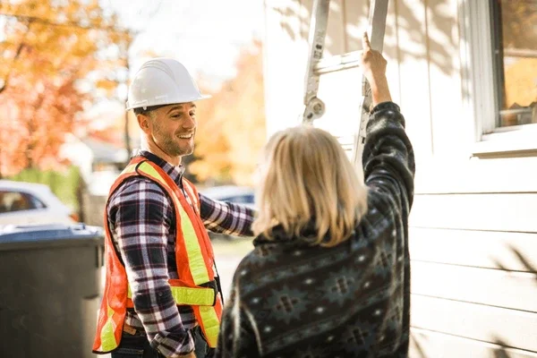 A man in a hard hat and safety vest smiling and talking to a woman holding a ladder against a house during daytime.