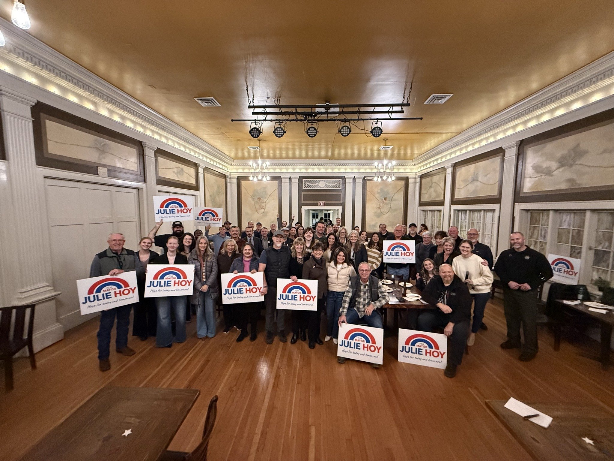 A large group of people gathered in a decorated banquet hall, holding signs that read "Julie Hoy" and "Hope for today and tomorrow." The attendees are smiling, and the hall has ornate molding and lighting on the ceiling.