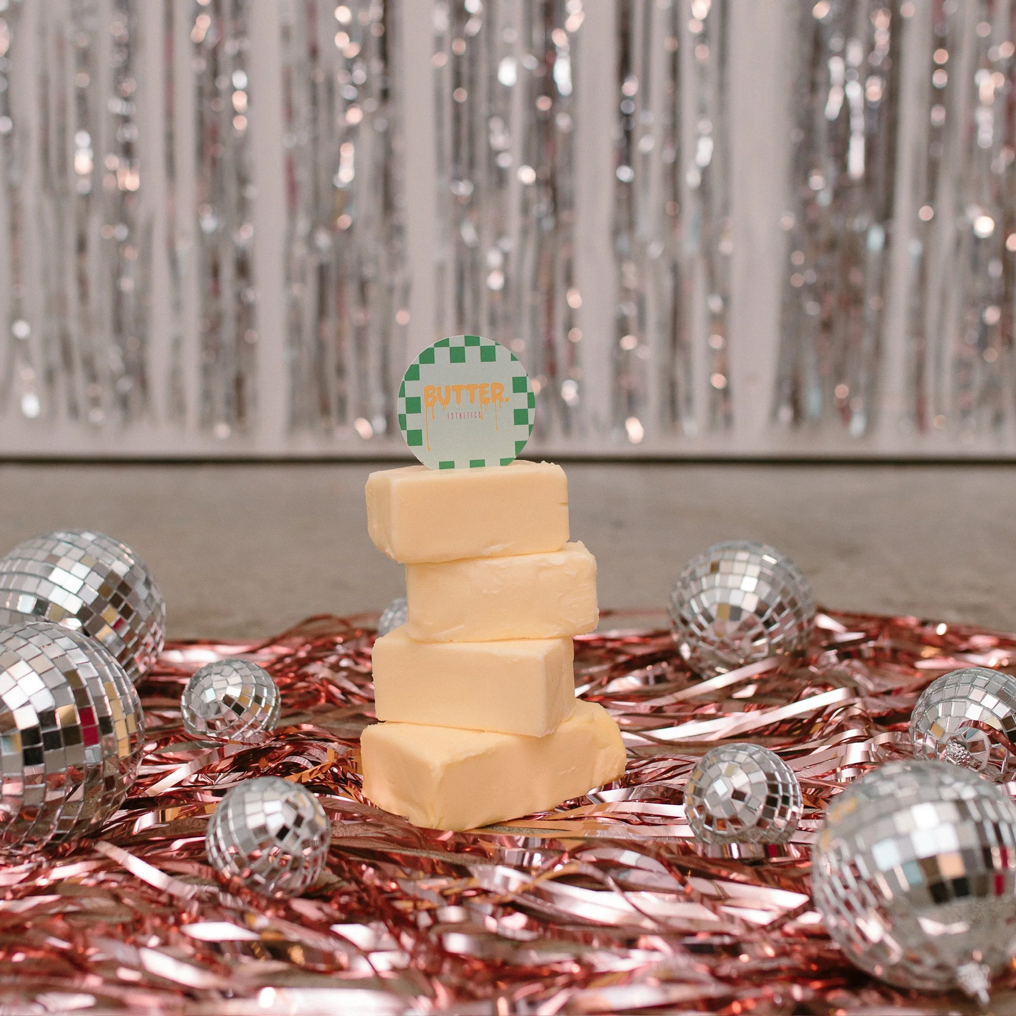 A display of various disco balls on a surface with a backdrop of shiny silver tinsel, featuring a small sign that reads 'Butter' in orange text with a checkerboard pattern border.