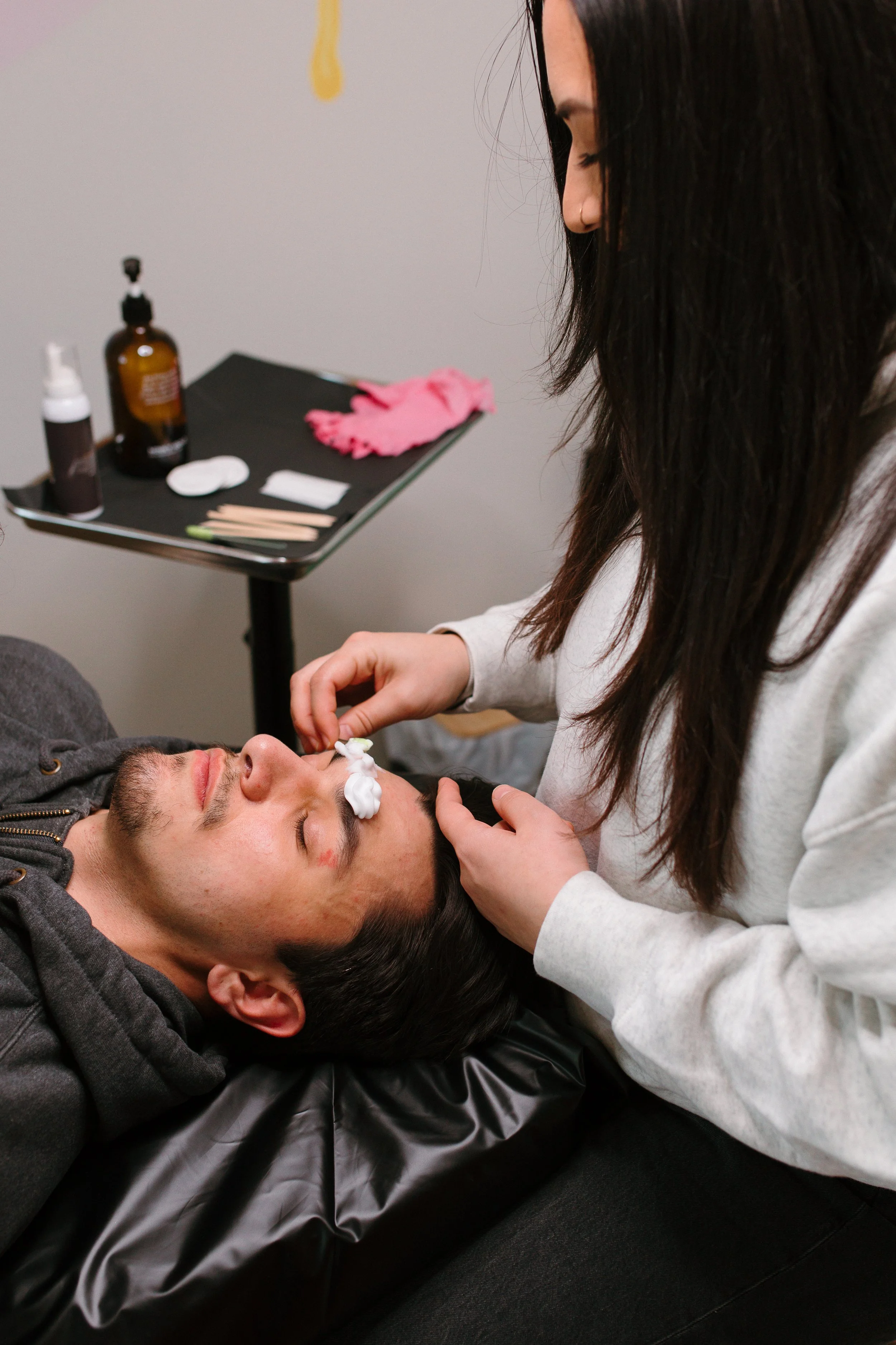 A woman performing a brow wax service on a man lying down with her hand on his face. Various skincare products and tools are on a nearby table.