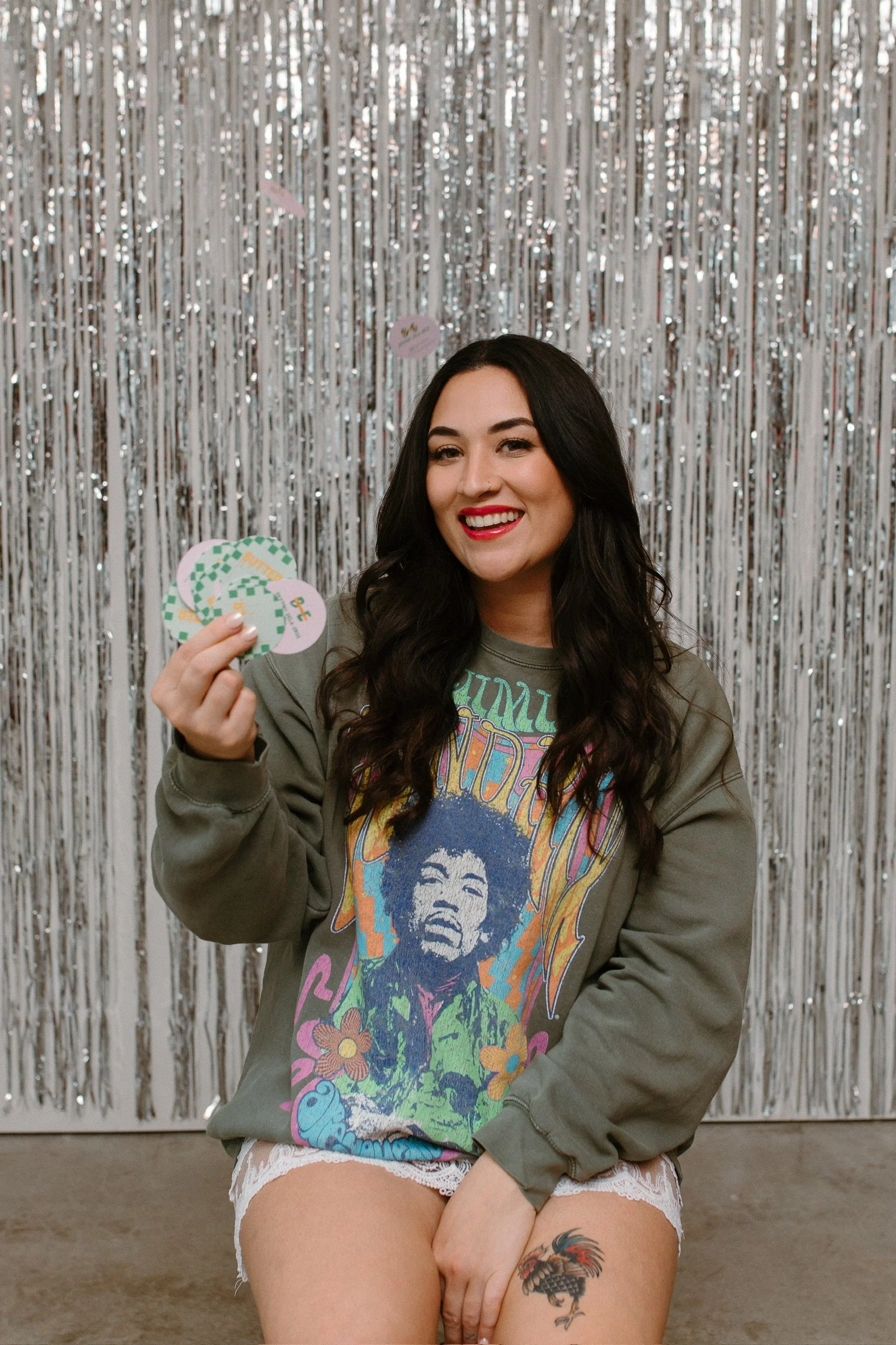 A woman with long dark hair and red lipstick is sitting in front of a silver tinsel backdrop. She is wearing a graphic t-shirt featuring Jimi Hendrix and a gray jacket. She is holding some cards or paper in her hand and smiling at the camera. She also has a tattoo of a rooster on her thigh.