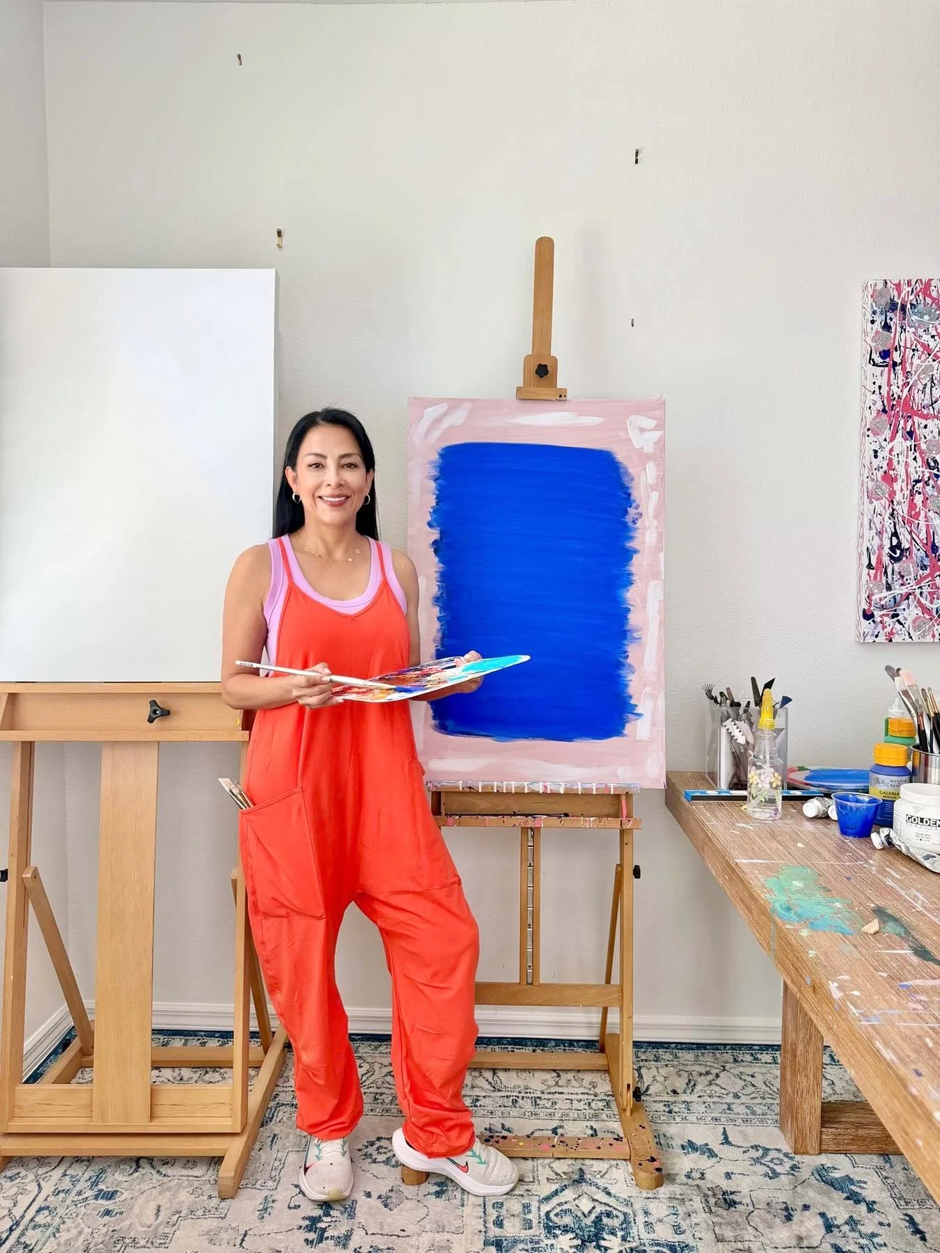 Artist Patricia Edelman in her studio — wearing a pink and orange outfit standing in art studio next to an abstract painting on easel, holding a paint palette and brush. Art supplies on table beside her.