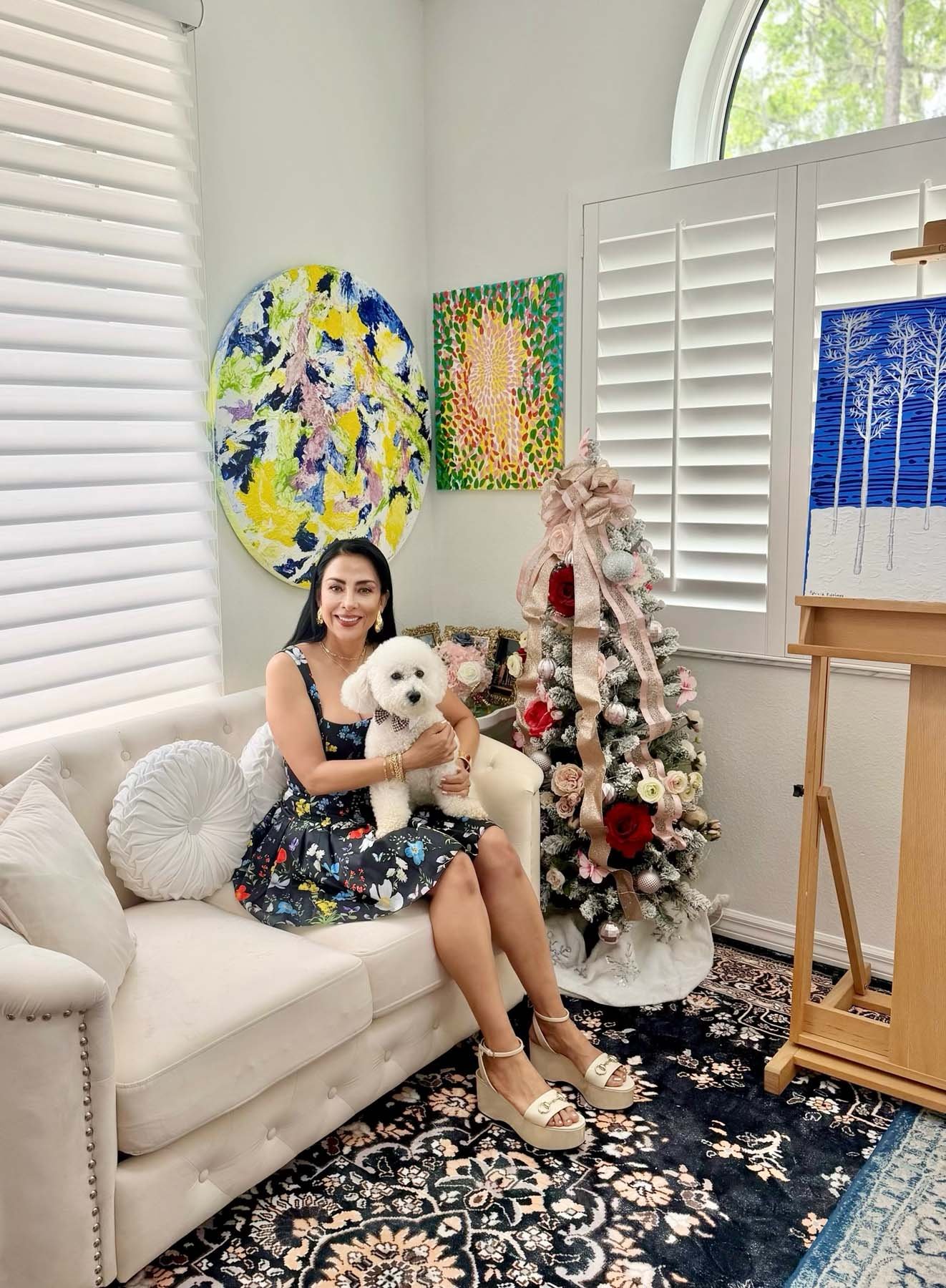 Artist Patricia Edelman — with black hair, wearing a floral dress and beige platform shoes, sitting on a white sofa, holding a white poodle dog, in a decorated living room with Christmas tree, colorful paintings, and a window with white shutters.