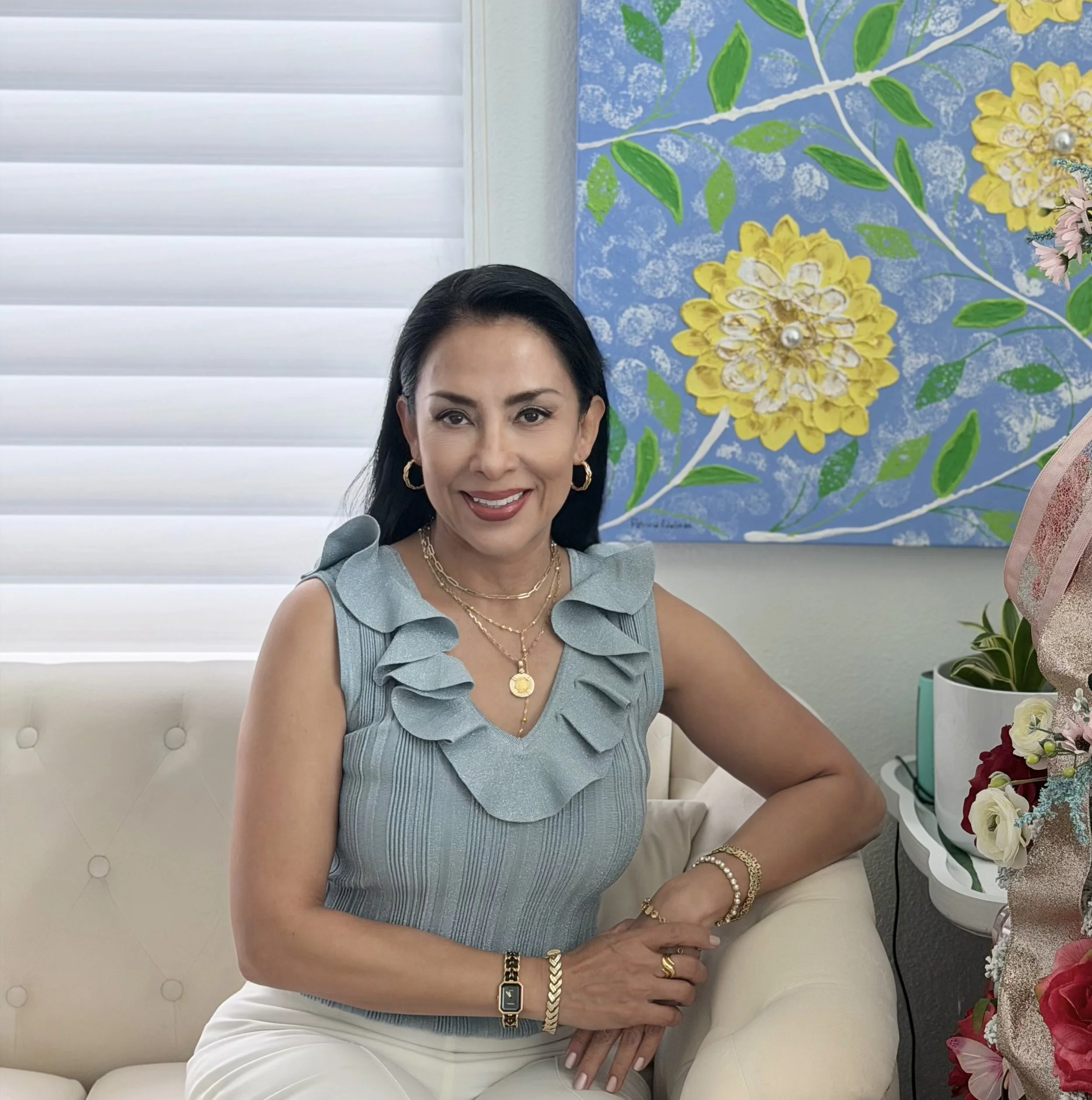 Artist Patricia Edelman — smiling, wearing gold jewelry, a light blue ruffled sleeveless top, and cream-colored pants. She is sitting on a white tufted chair, with a floral background featuring yellow and pink flowers and green leaves.