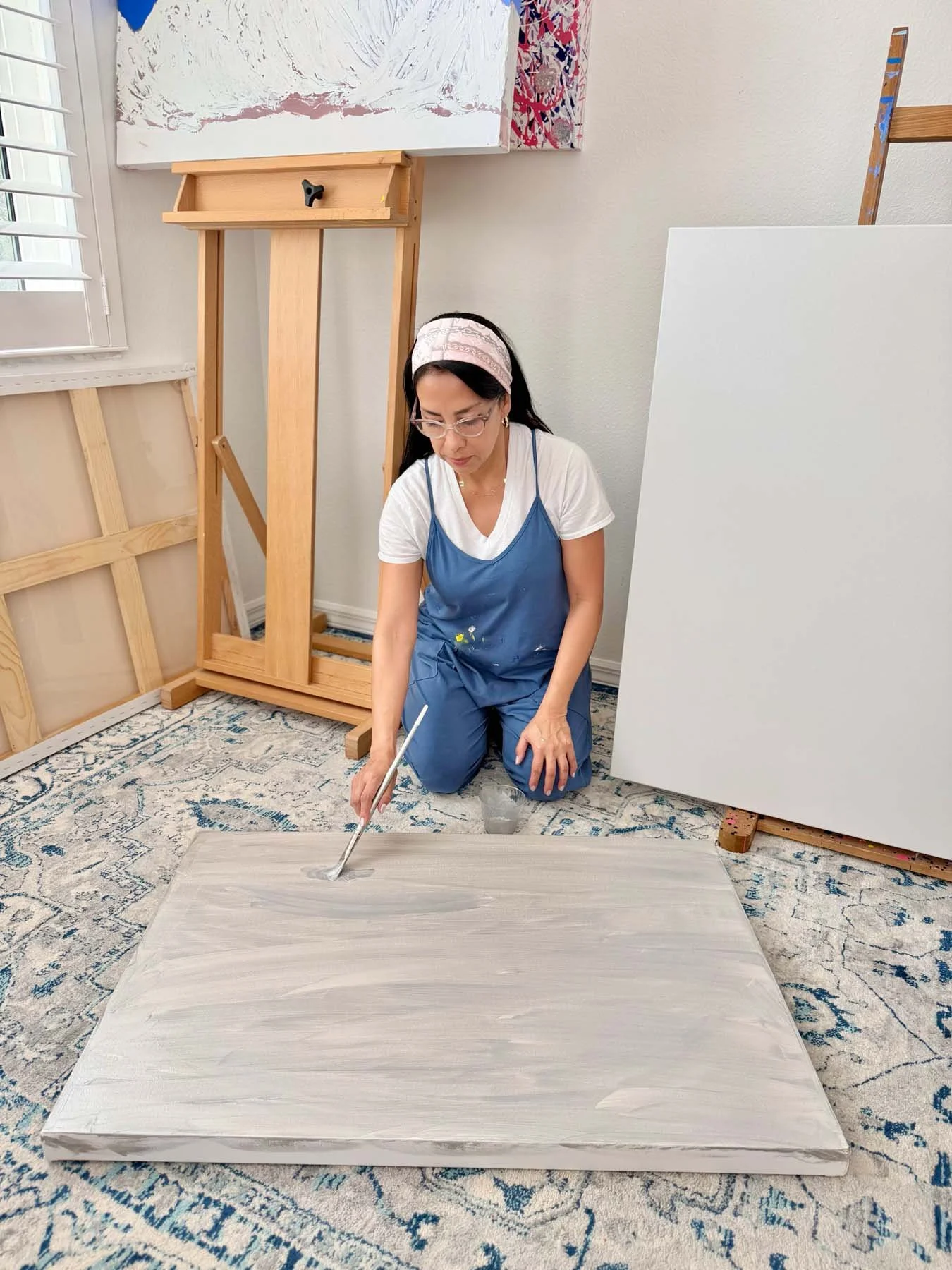 Artist Patricia Edelman in her studio, kneeling on a patterned rug, painting a large canvas in an art studio, surrounded by other unpainted canvases and art supplies.