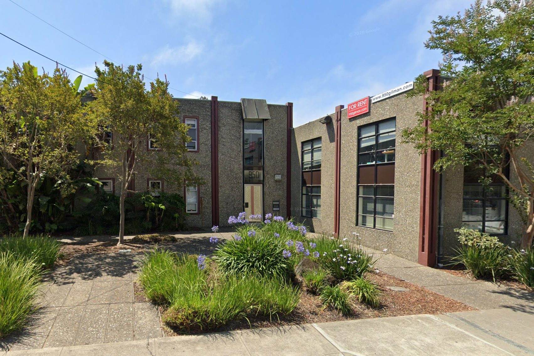 A two-story commercial building with large windows, modern concrete exterior, and a sign indicating it is for rent at 950. There are trees, plants, and flowers in front of the building and a sidewalk.