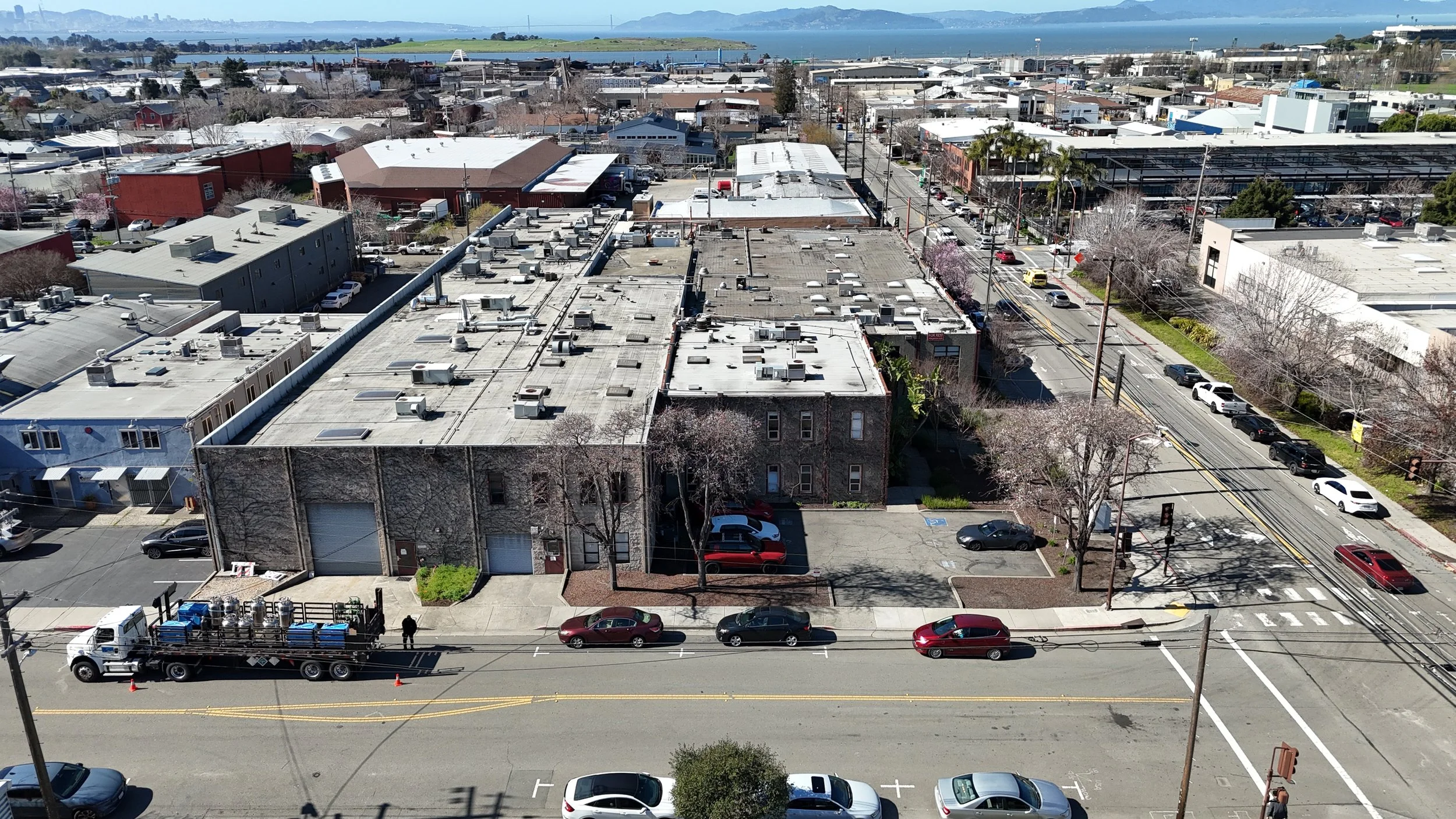 Aerial cityscape view showing rooftops, streets with parked and moving cars, palm trees, and a distant body of water with hills or islands.