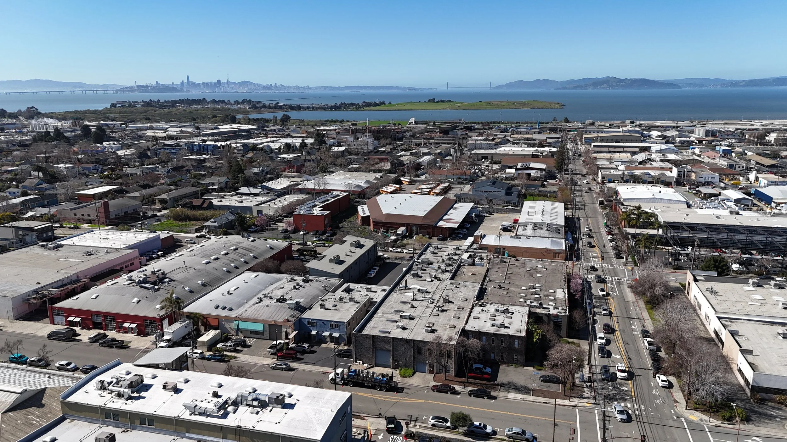 Aerial view of a city neighborhood with commercial and residential buildings, streets with cars, near a body of water with islands and the skyline of San Francisco in the background.