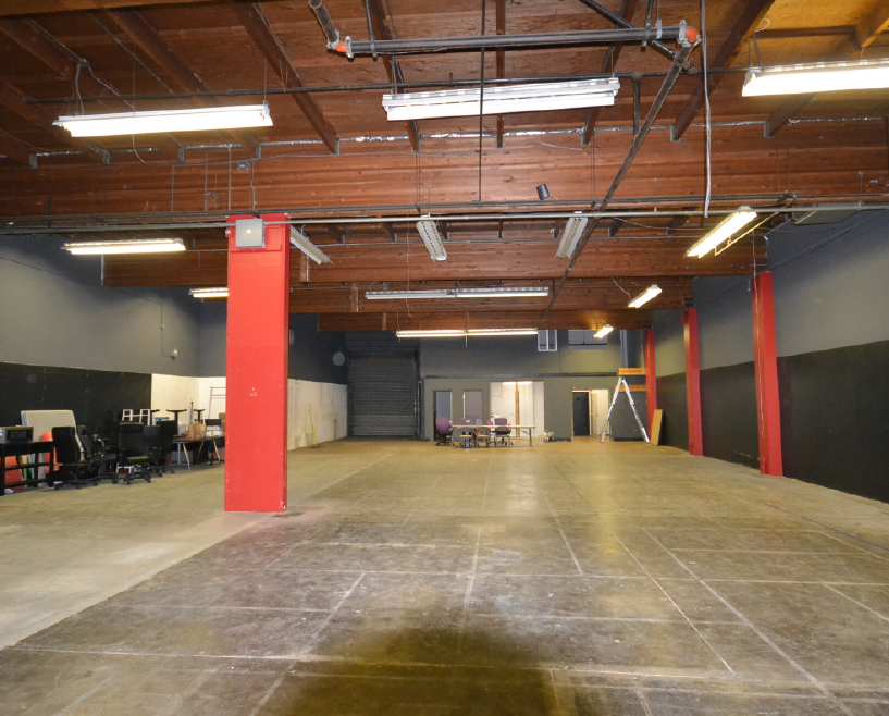 Empty industrial or commercial space with fluorescent lighting, exposed wooden ceiling, concrete floors, and some chairs and tables at the back.