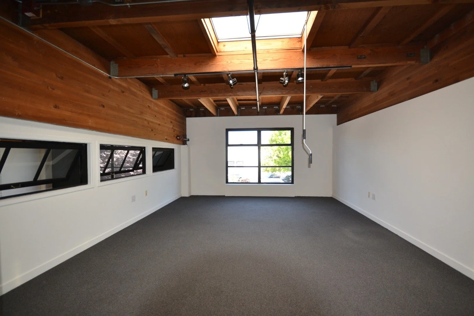 Empty room with white walls, gray carpet, wooden ceiling with exposed beams, skylight and window, and multiple electrical outlets.