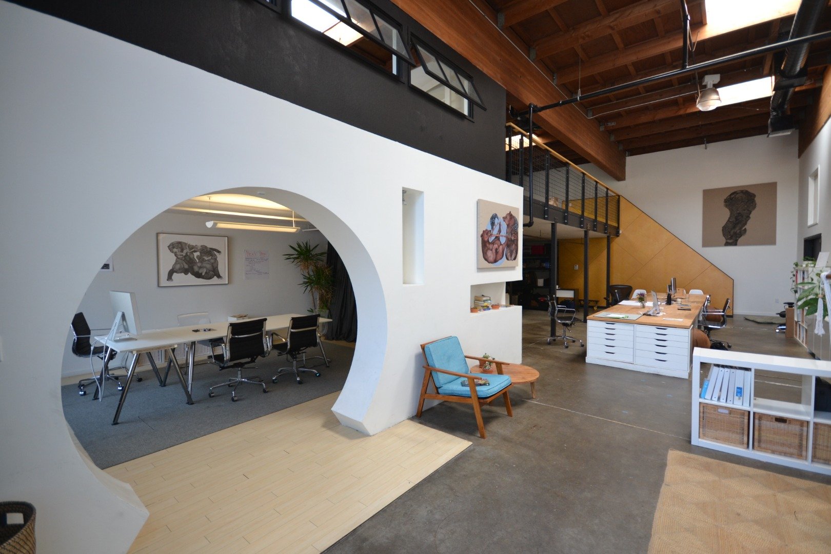 Modern open-concept office with a white wall and wooden ceiling, featuring a lounge area with a blue armchair and small table, a conference room with a curved wall and black chairs, and workstations with computers and art on the walls.