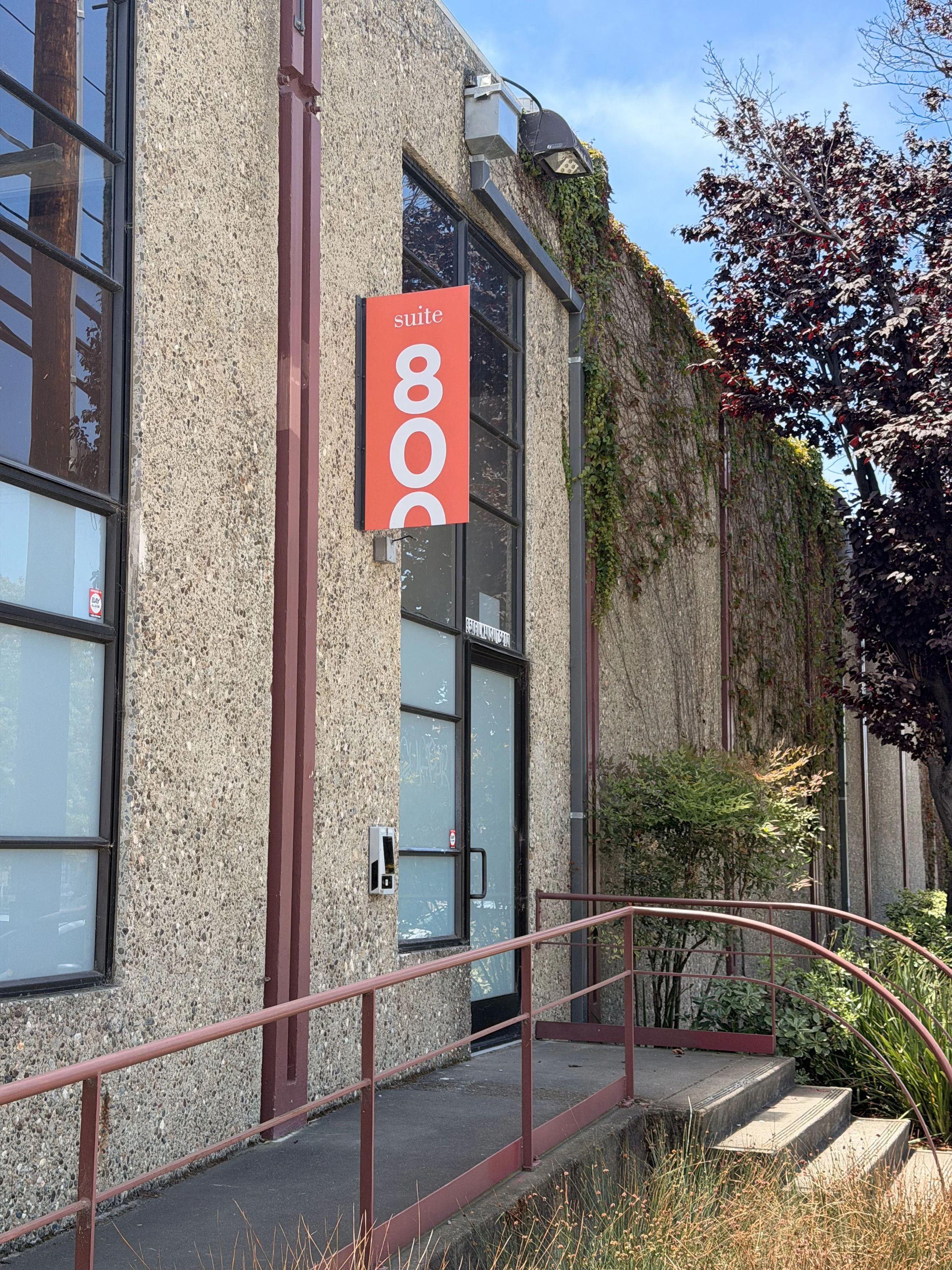 A building with a textured concrete wall and large windows. A red sign on the wall reads 'suite 800'. There is a wheelchair accessible ramp with a red metal railing leading up to the door, surrounded by plants and trees with autumn-colored leaves.