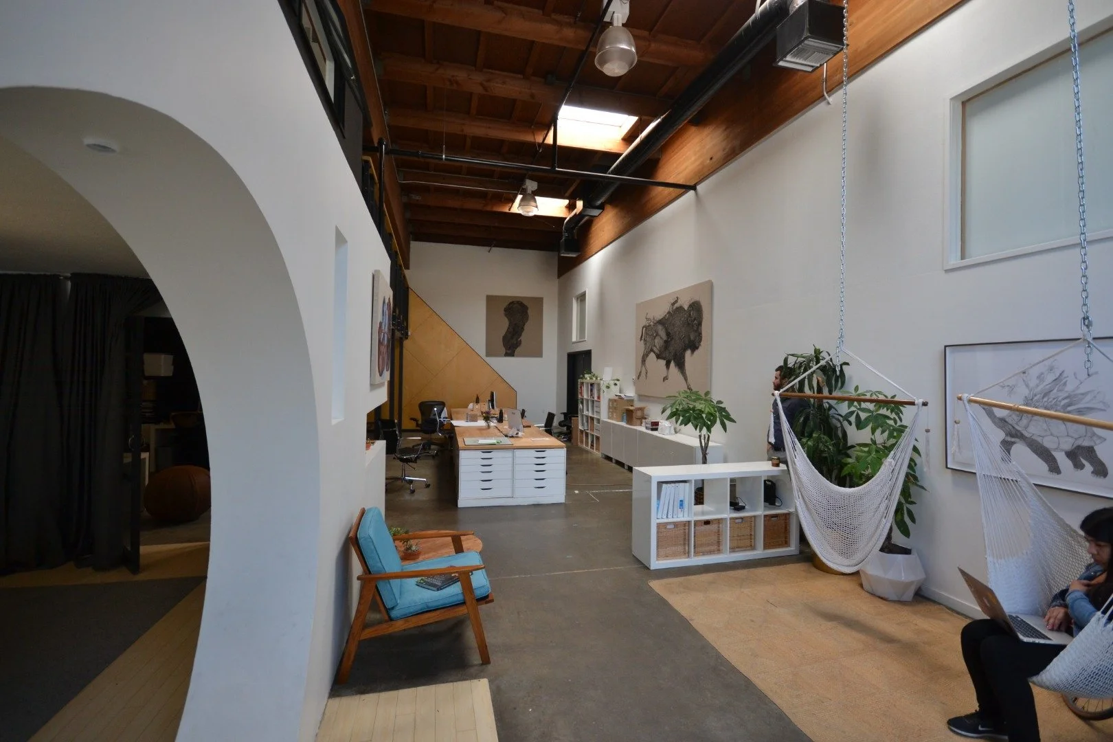 Interior of a modern office with high wooden ceiling, white walls, wall art of animals, large plants, hammock chair, and two people working on laptops.