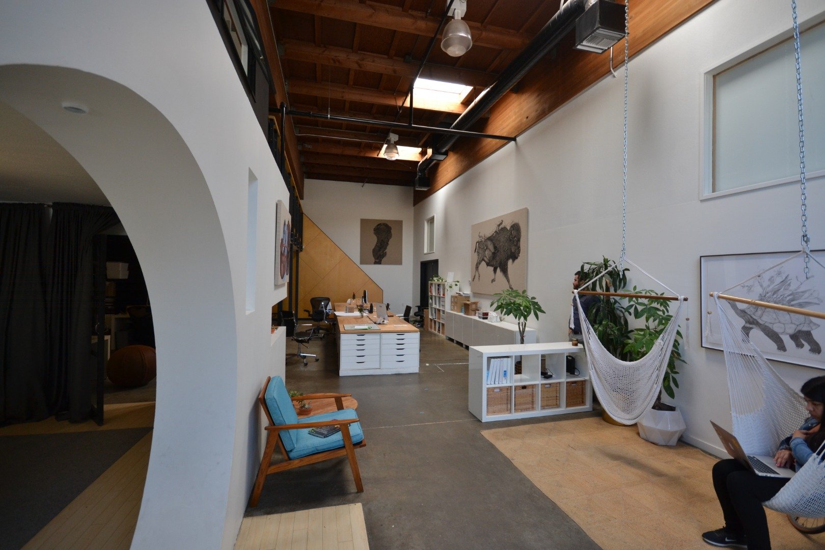 Open-plan office space with high wooden ceiling, white walls, and artwork of buffaloes. Seating includes a wooden chair with blue cushions and a person working on a laptop. The space has plants, a hammock, and office desks with chairs.