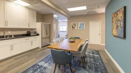 Bright dining area with a wooden table, three blue upholstered chairs, and a modern kitchen with white cabinets and stainless steel appliances.