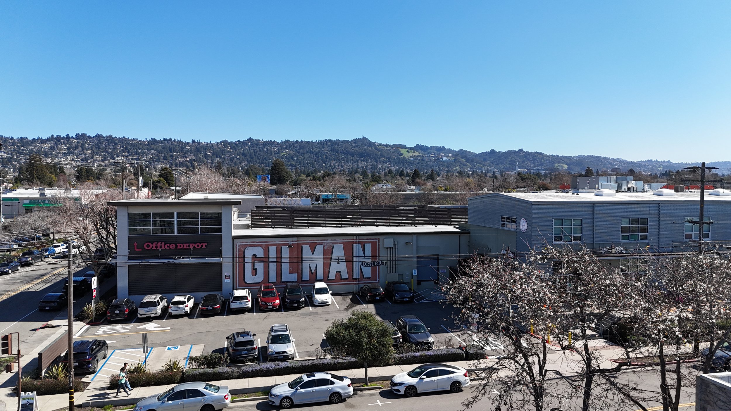 Cityscape view with commercial buildings, parking lot with cars, and rolling hills in the background under a clear blue sky.