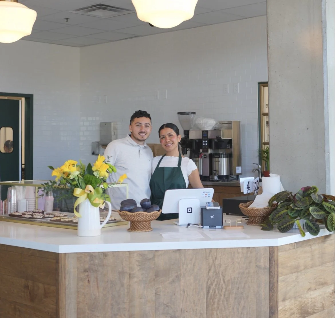 Smiling man and woman behind the counter of a cafe with a white countertop, yellow flowers, avocados, and a cashier terminal.
