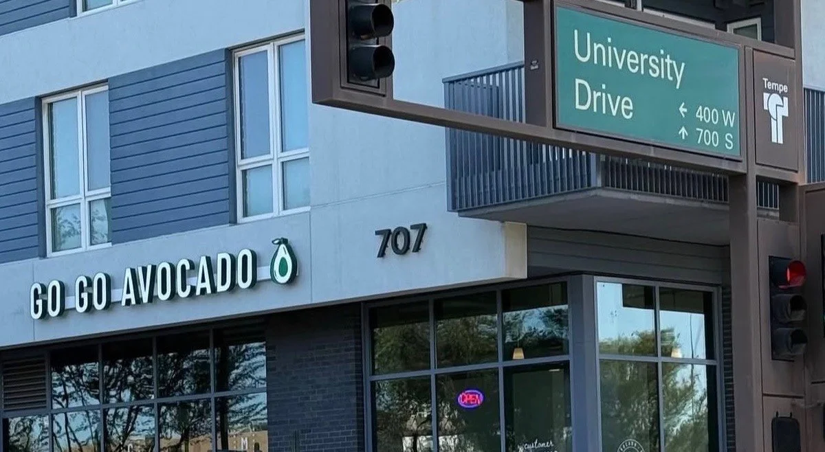 The image shows a storefront called 'Go Go Avocado' at 707 on a street named University Drive, with a green sign overhead indicating nearby street intersections. The store has large glass windows and an illuminated 'OPEN' sign inside.