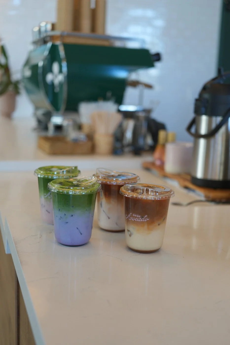 Four cups of iced beverages with lids on a light-colored countertop, two with green topping, and two with chocolate topping, in a cafe setting.