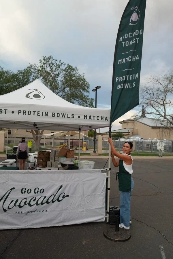 A woman standing next to a promotional booth for avocado toast, matcha, and protein bowls at an outdoor event, holding a tall flag with similar branding.