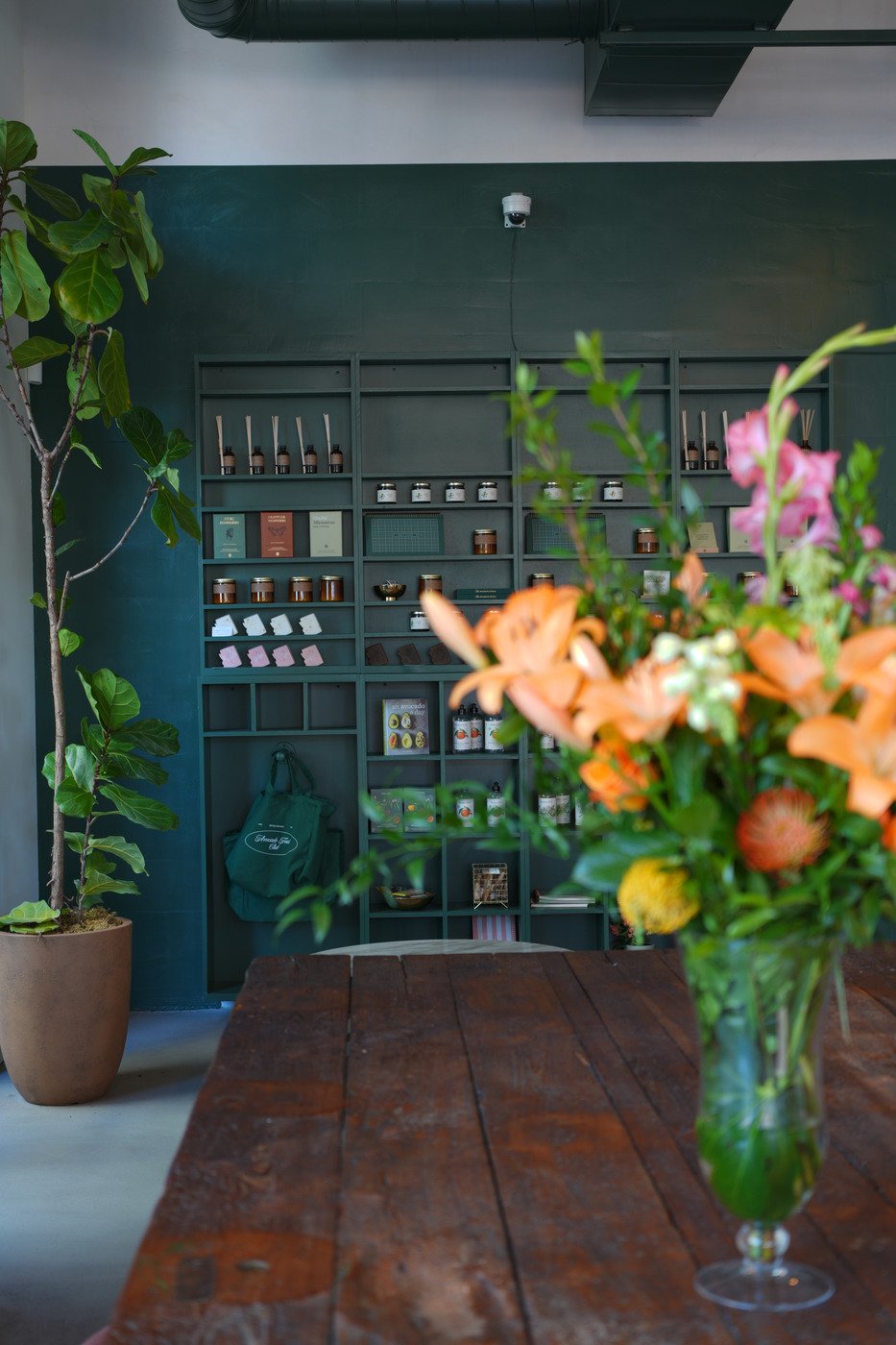 Interior of a store or cafe with a dark green wall shelving unit holding jars, books, and small items, with a large potted plant on the left and a table with a colorful flower bouquet in the foreground.