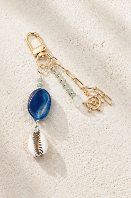 A decorative keychain with beads, a blue stone, and a cowrie shell charm, lying on a textured beige surface.