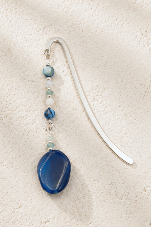 A silver bookmark with a beaded blue and white ornament hanging from it, resting on a textured white surface.