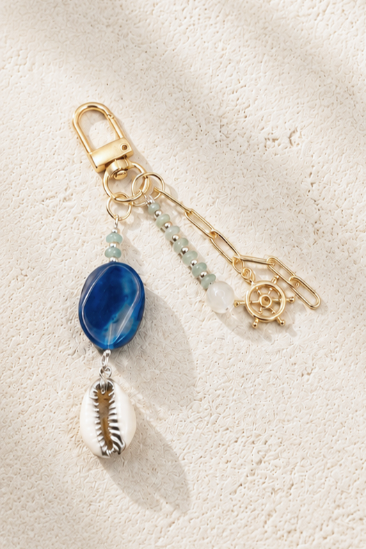 Gold safety pin keychain with blue bead, white bead, small light blue beads, and cowrie shell charms attached, lying on textured light-colored fabric.